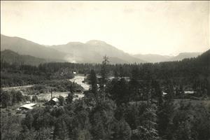 Wooden buildings in the trees along the shores of a winding river with mountains in the background. Image is sepia. 